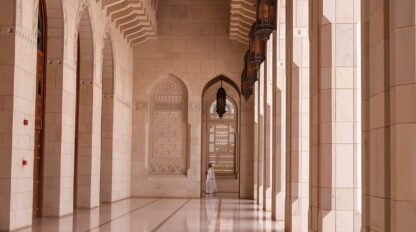 A person in white stands in the corridor of a grand mosque with tall arches, columns, and intricate wall designs, echoing the serene beauty of Oman's mountains.